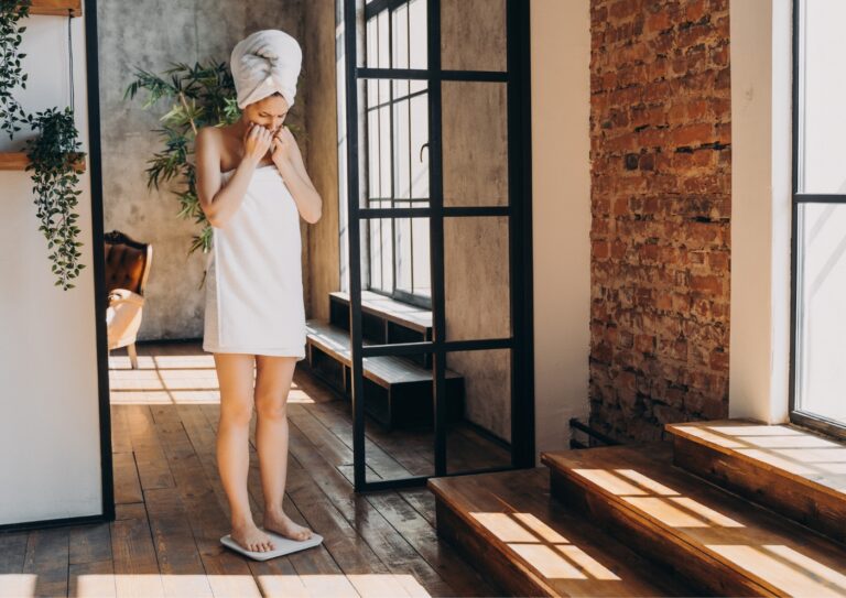 40 plus vrouw komt net onder douche vandaan en heeft een handdoek gewikkeld om haar lichaam en een om haar hoofd. Ze staat haar handen in haar mond op een weegschaal en kijkt met een bezorgde blik naar het resultaat