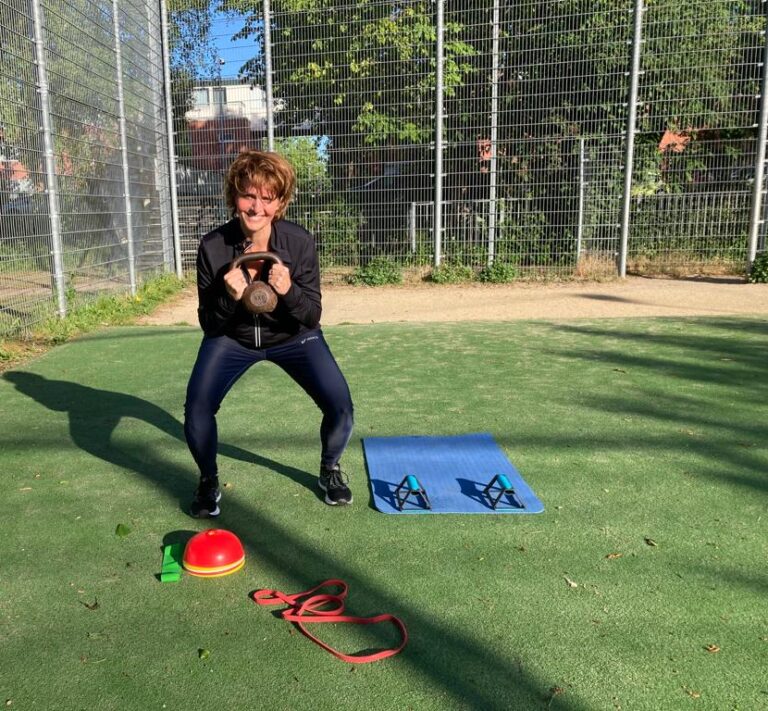 Fitte vrolijke vrouw traint buiten op een voetbalveldje op kunstgras. Houdt een kettlebell voor zich vast en lacht in de camera